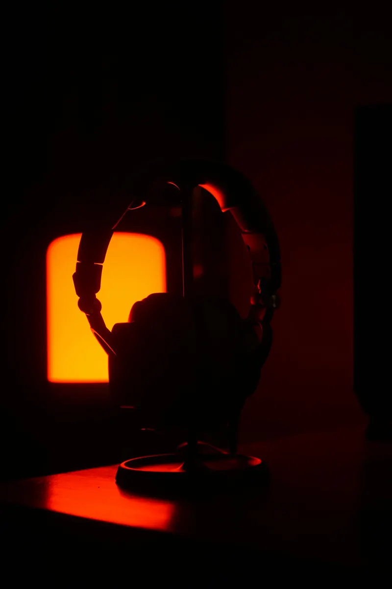 Over-ear headphones on a desk stand, silhouetted against a warm amber lamp in a dark room
