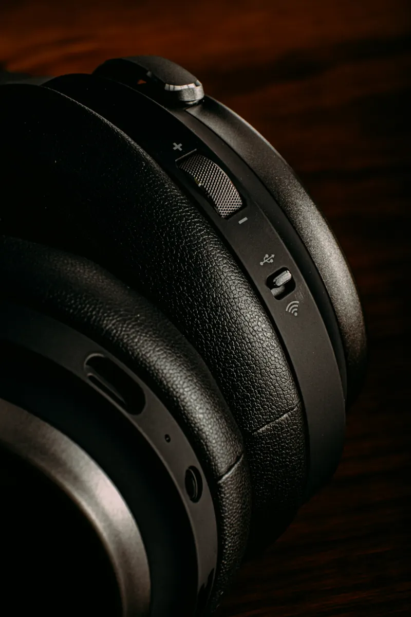 Ear cup of a black wireless headphone with volume rocker and switch, resting on dark wood